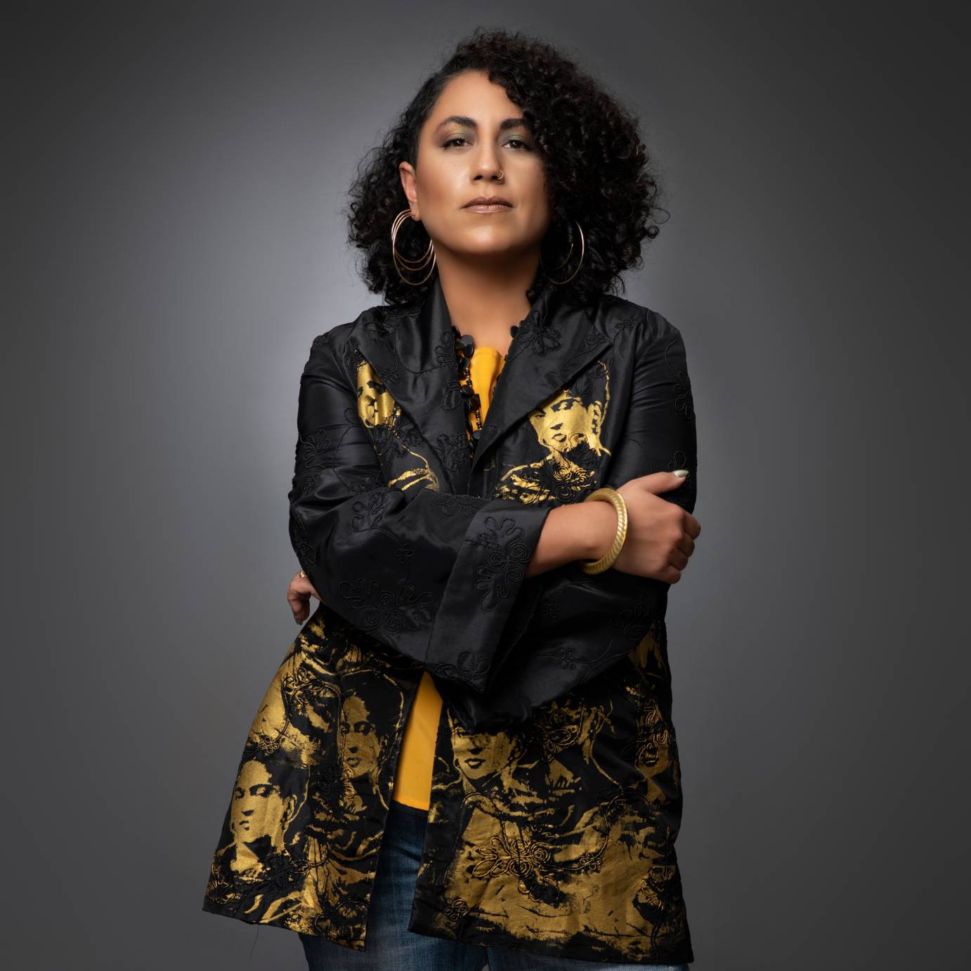 photo of a light brown skinned woman composer Taina Asili staring at us. She wears large hoop earrings a bright yellow shirt and blue jeans topped off by a loose fitting black jacket decorated with gold images of Frida Kahlo.